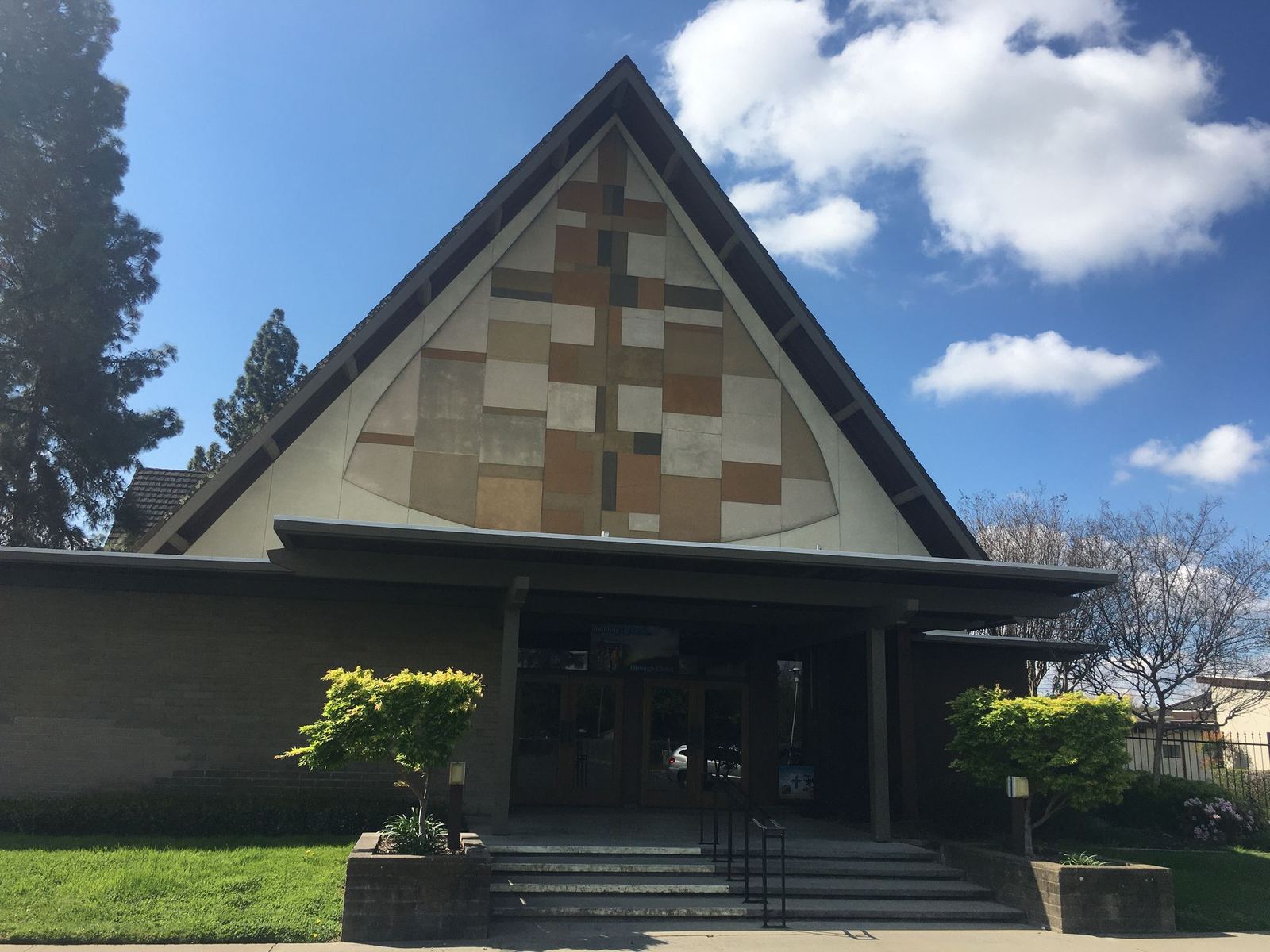 The front of the church on a clear day: a steep A-frame roof over a mosaic cross, with glass doors beneath a low entrance canopy