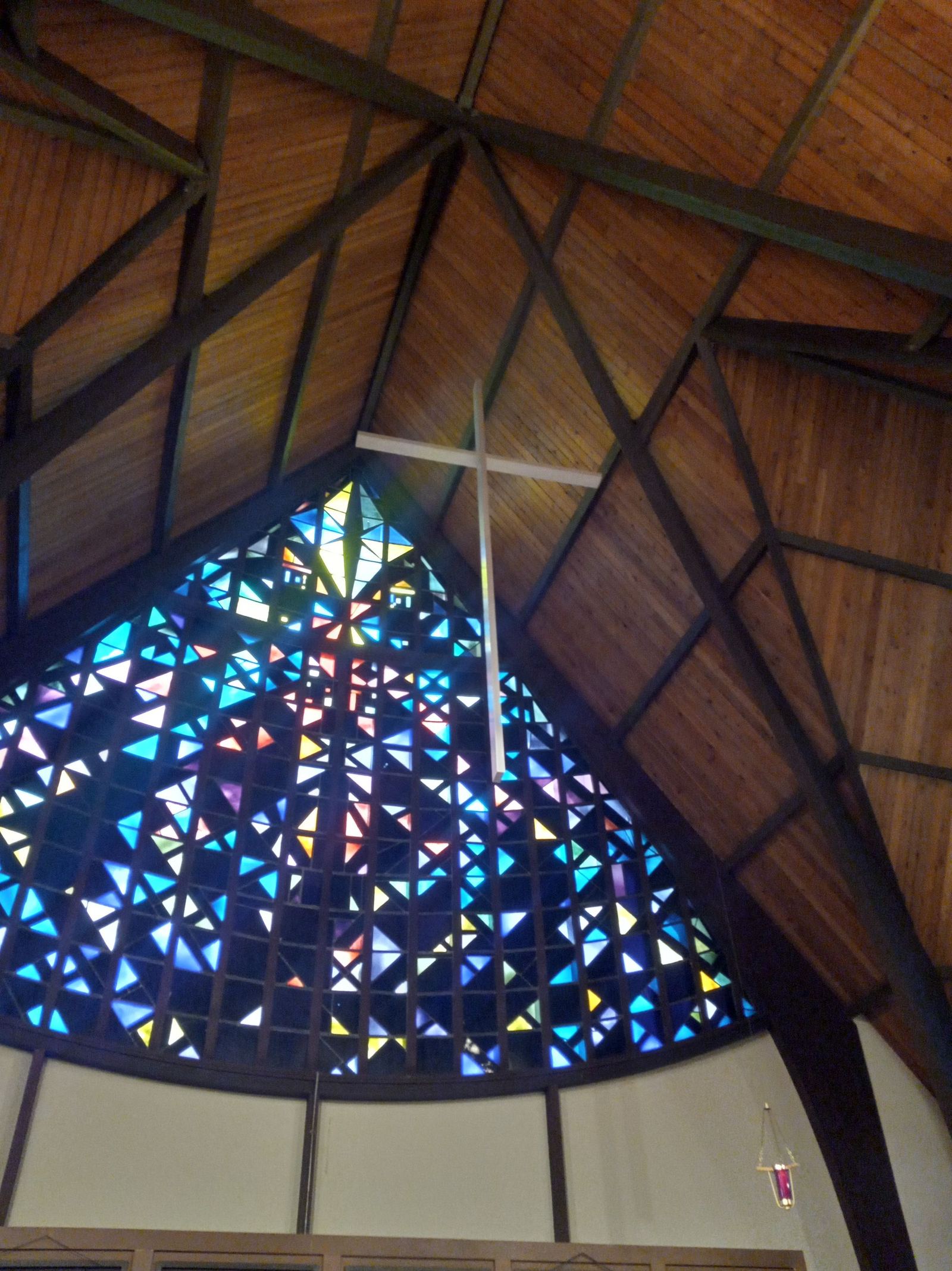 The sanctuary's stained glass window, a mosaic of small colored triangles, with a plain white cross hanging in front of it