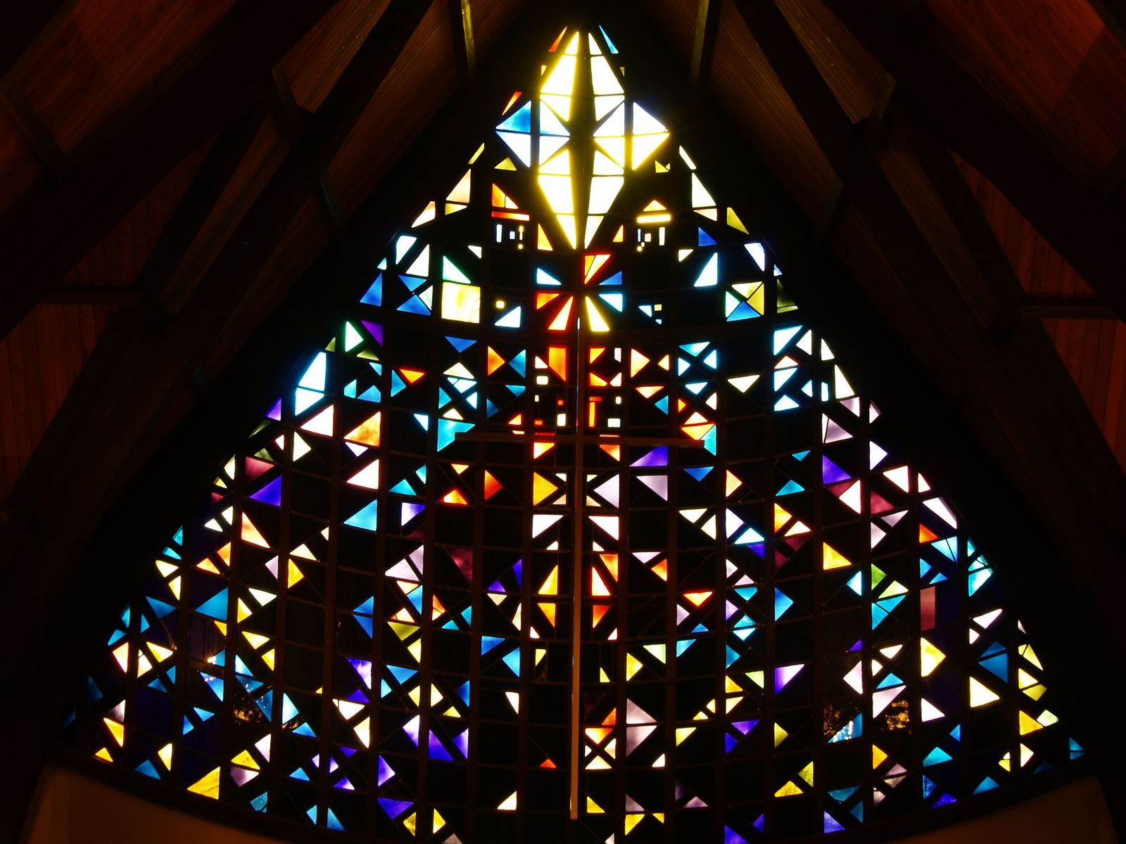 The stained glass window lit from behind in a darkened nave: hundreds of small colored triangles around a cross