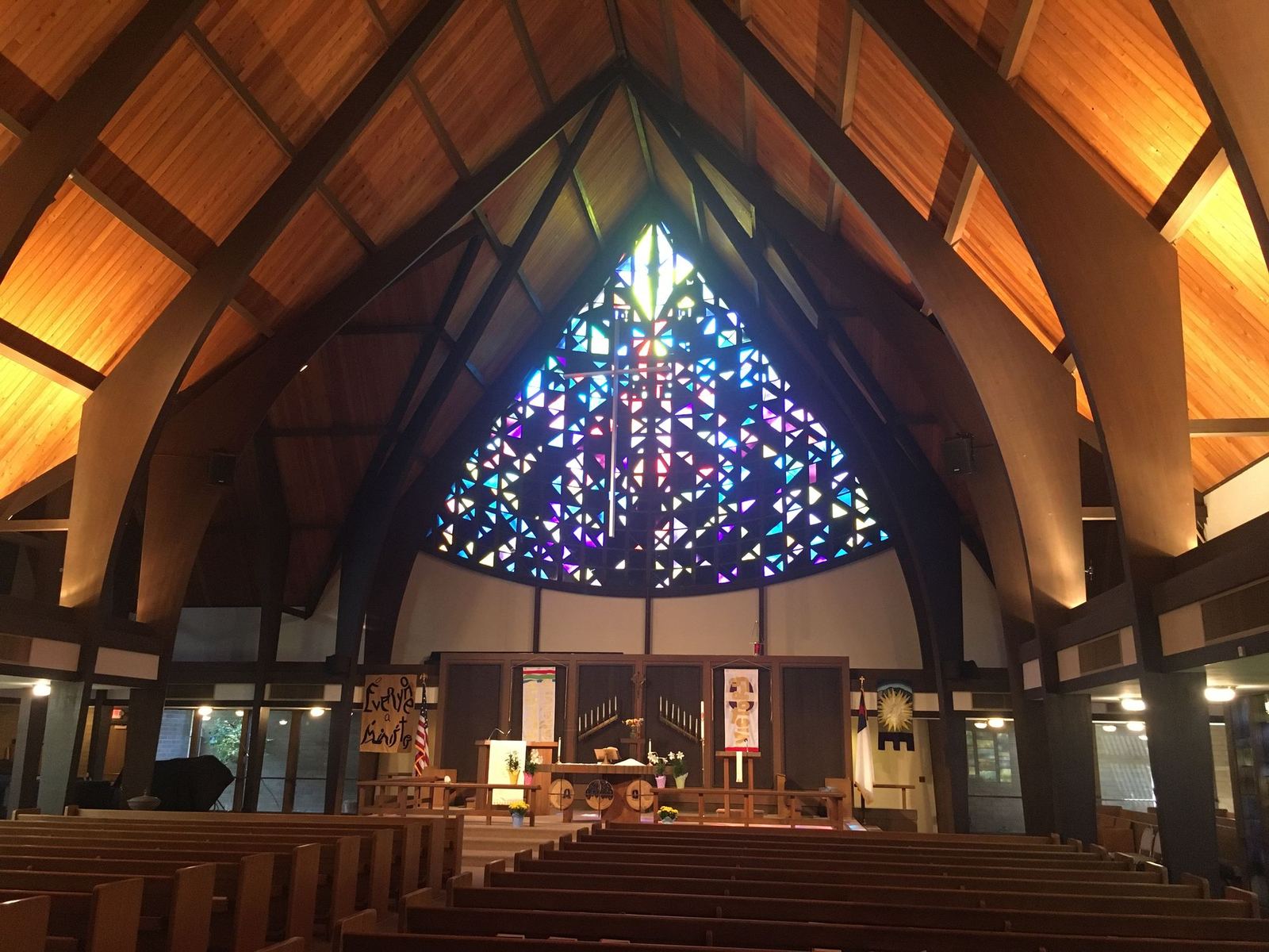 The chancel seen down the center aisle, with the stained glass window above the altar, banners on either side, and lilies at the foot of the altar