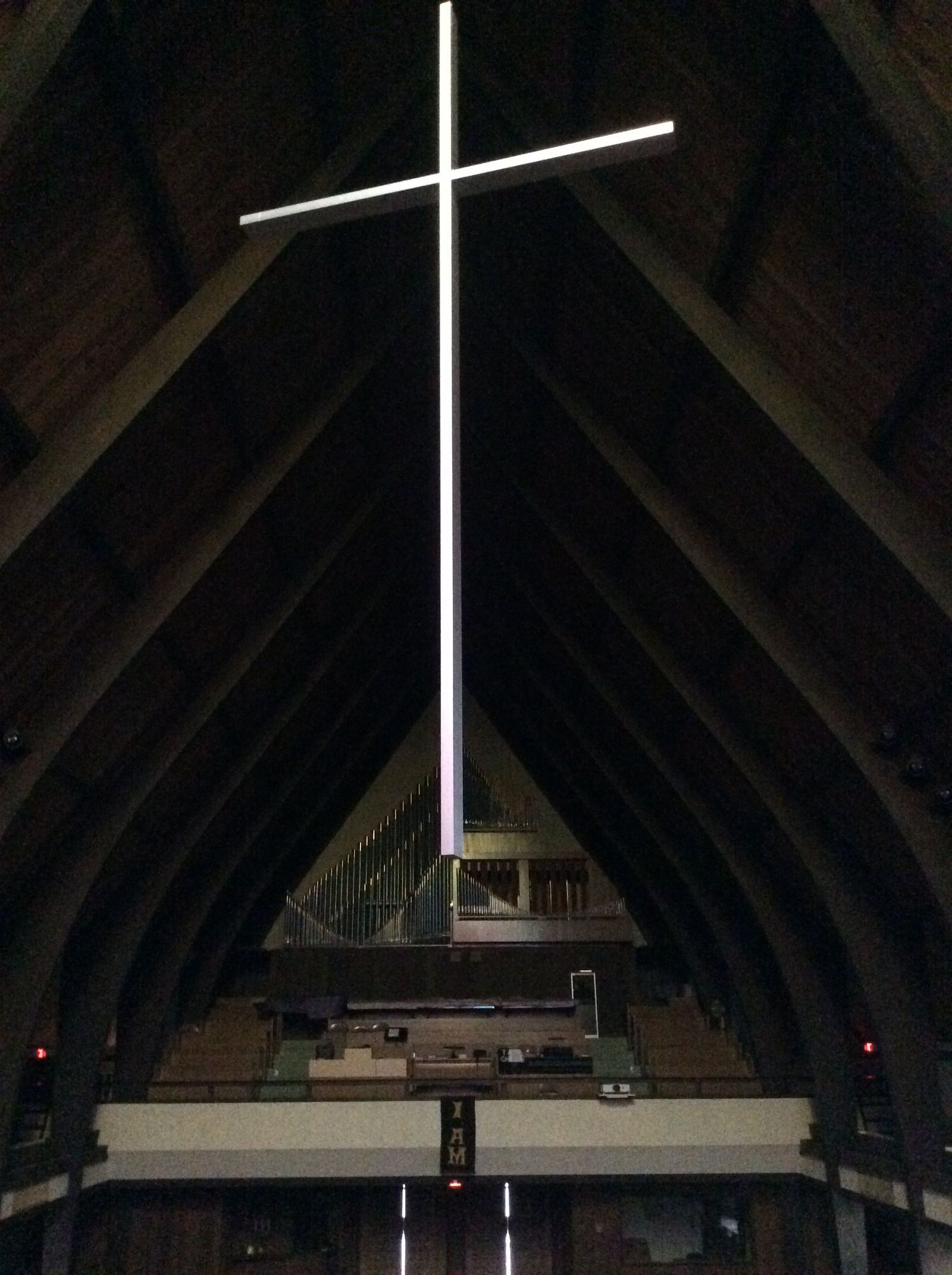 The white cross hanging from the sanctuary ceiling, lit against the dark timbered roof, with the organ pipes below
